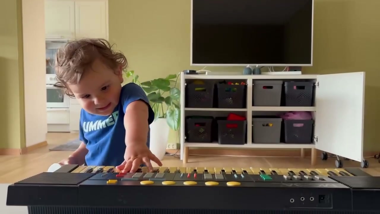 A curly-haired toddler in a blue "Summer" t-shirt reaching out to press the keys of a large old electronic keyboard in a living room
